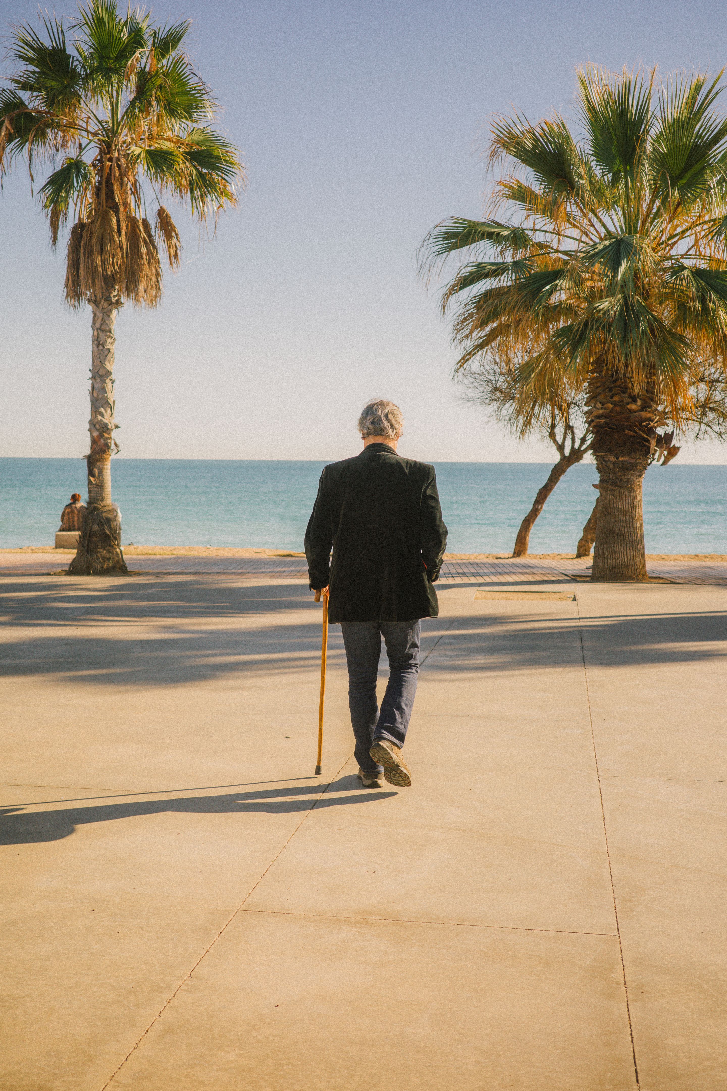 Foto de un hombre mayor de espaldas con un bastón que camina en dirección al mar, que está al fondo.