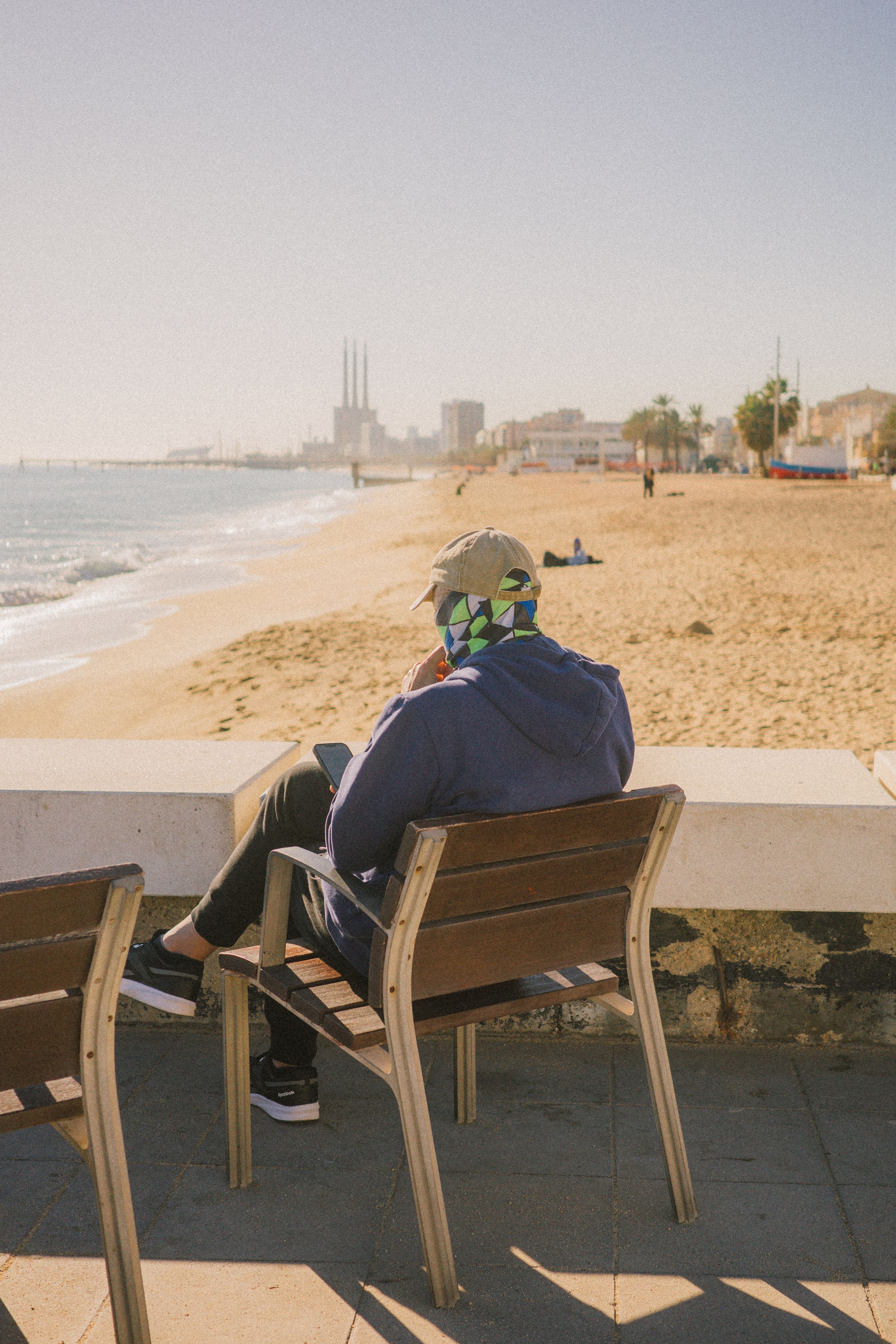 Una persona sentada en un banco mira a su teléfono. Lleva una gorra beige y un pasamontañas de colores. Al fondo se ve la playa y las Tres Chimeneas de Sant Adrià de Besòs.