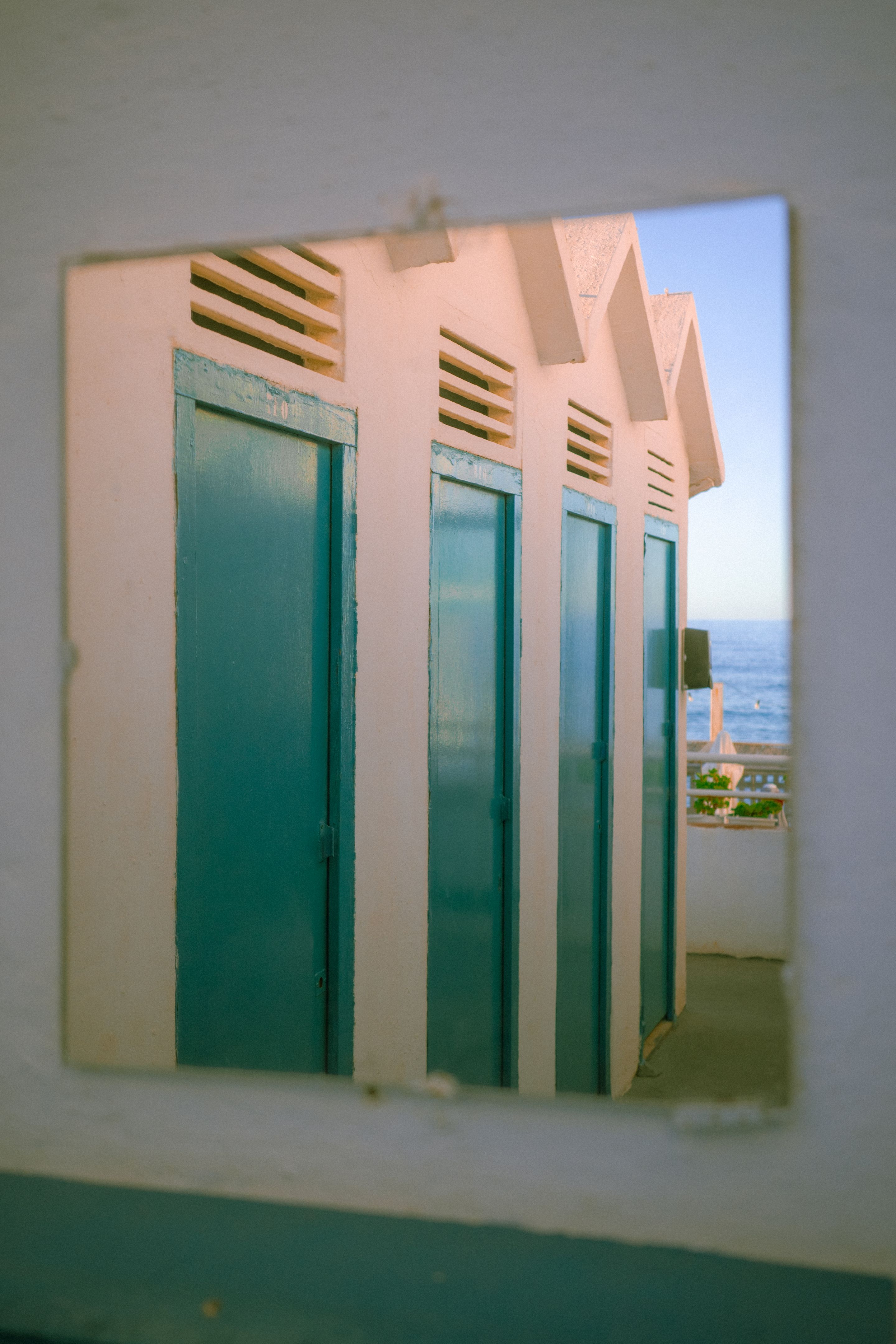 Foto de unas casetas de baño al atardecer. Las casetas se ven en el reflejo de un espejo. Al fondo de la imagen se aprecia el mar.