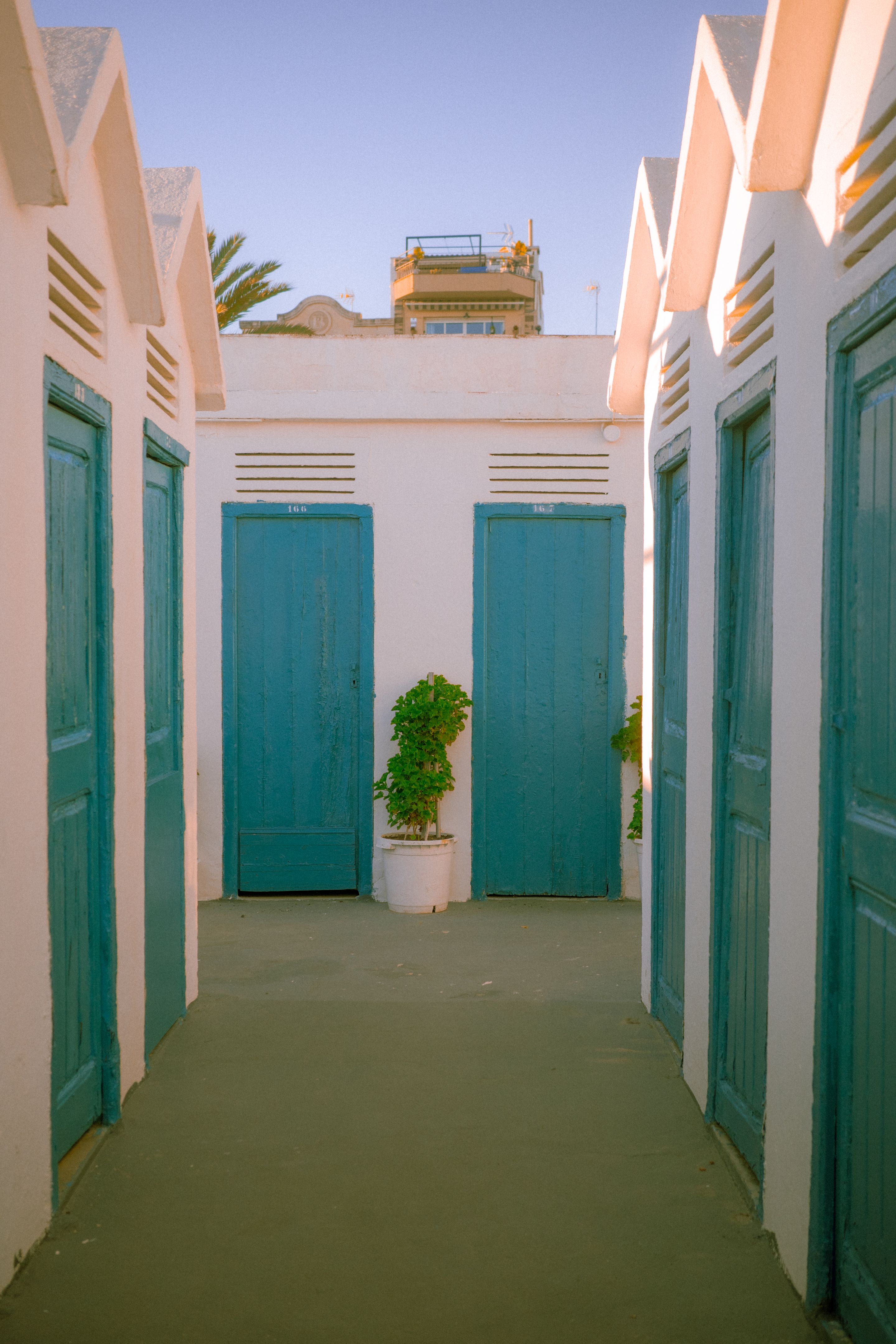 Foto de unas casetas de baño al atardecer. En la imagen se pueden ver las puertas azules de entrada. Al fondo de la imagen se ve parte de un edificio.