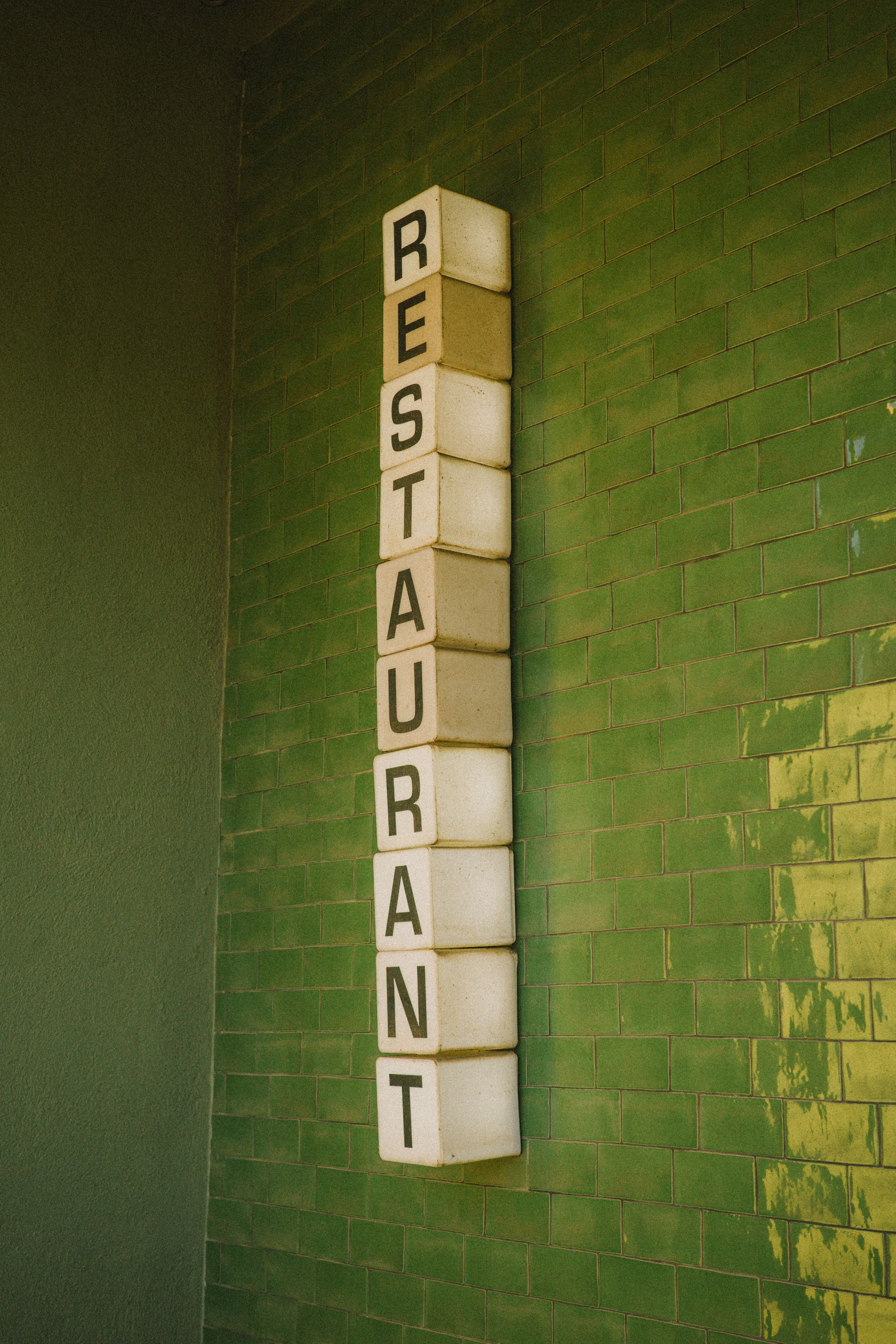 Foto de un letrero donde se puede leer la palabra 'restaurant' en vertical. Cada letra está dibujada sobre un cubo blanco. El letrero está colgado sobre una pared de ladrillo verde.