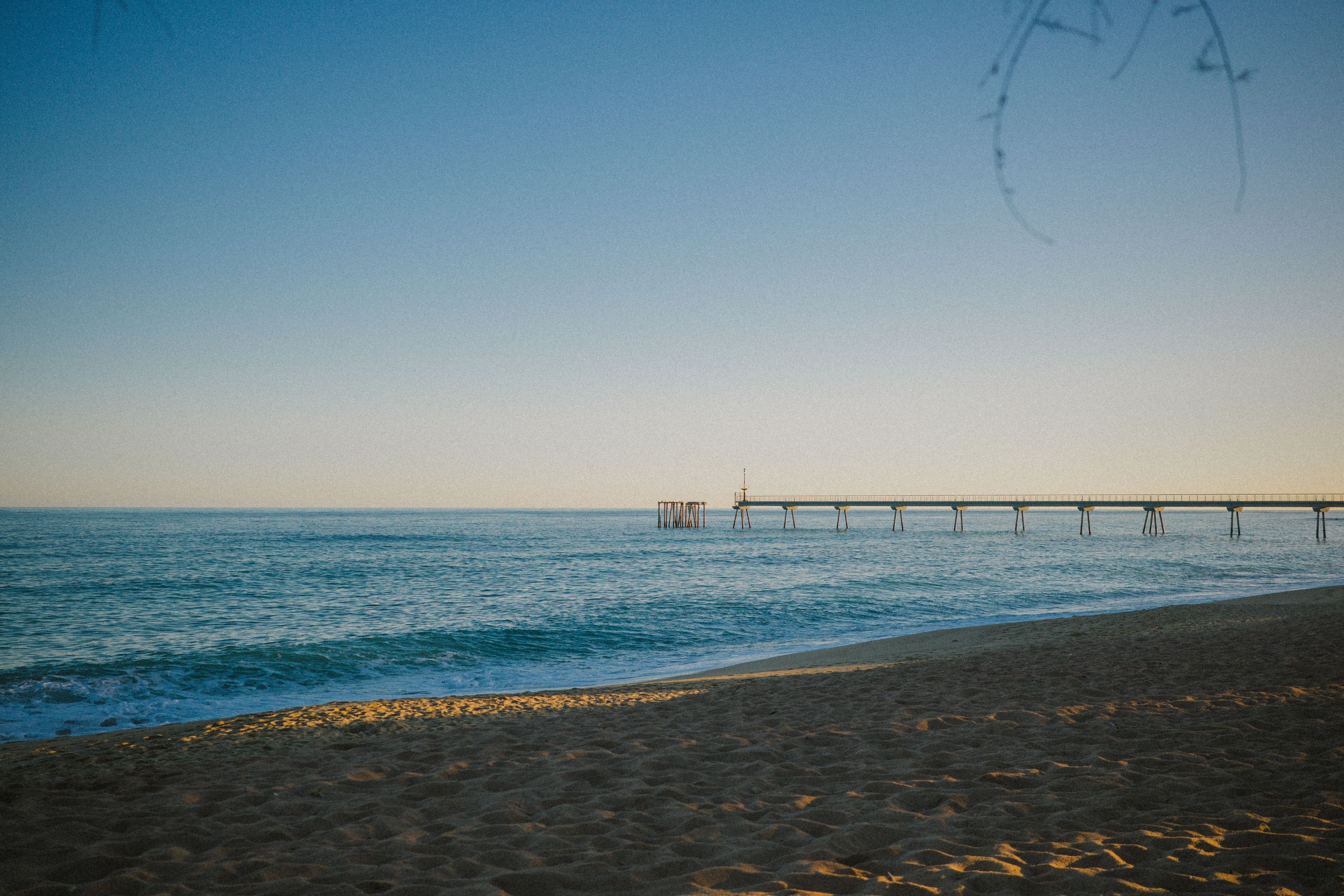 Foto de una playa y el mar al atardecer, sin nubes, y con una plataforma de madera al fondo.