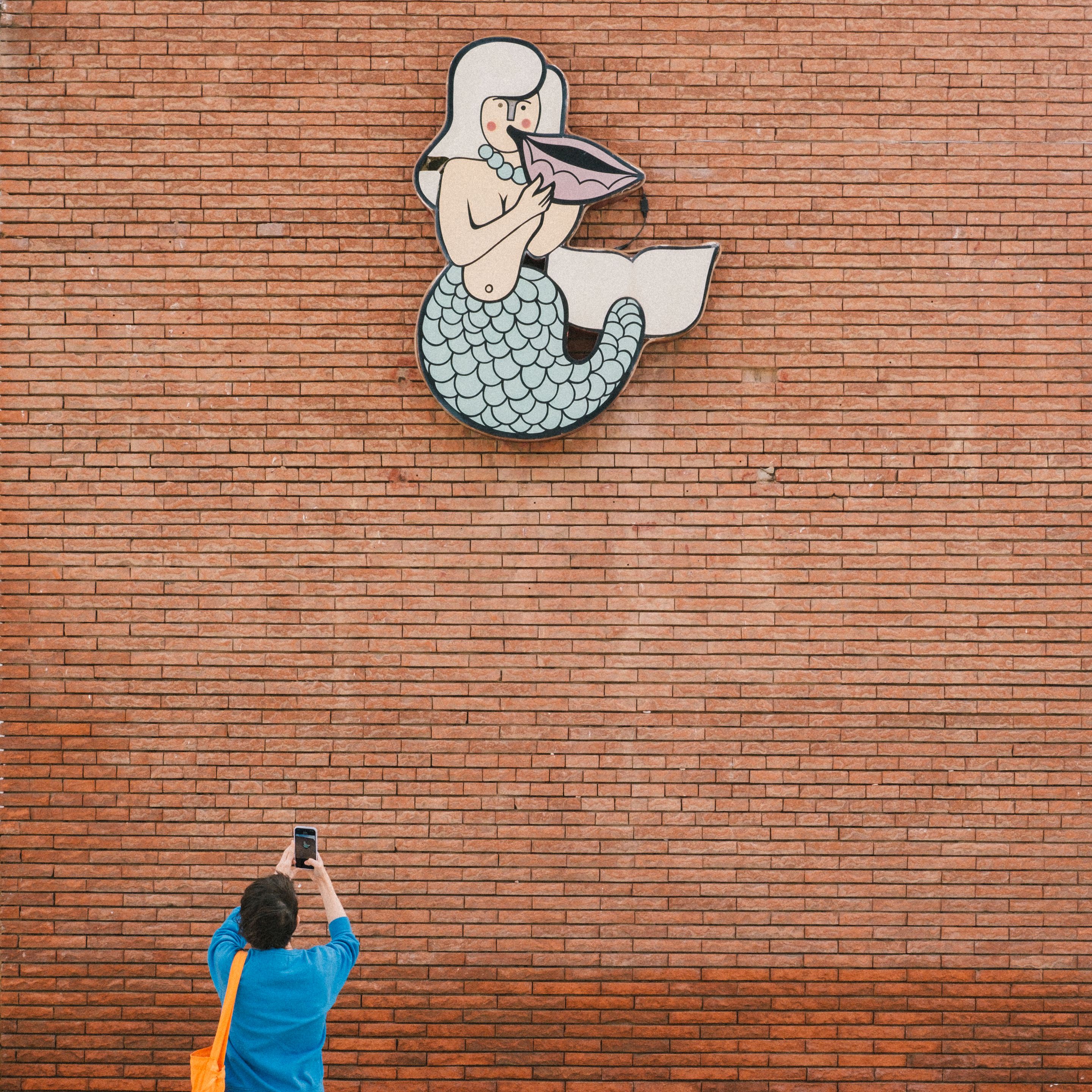 Foto de una pared y el dibujo de una sirena tocando una concha sobre una pared de ladrillo naranja. En la esquina inferior izquierda una chica de espaldas con camisa azul y un bolso naranja le hace una foto con el móvil.