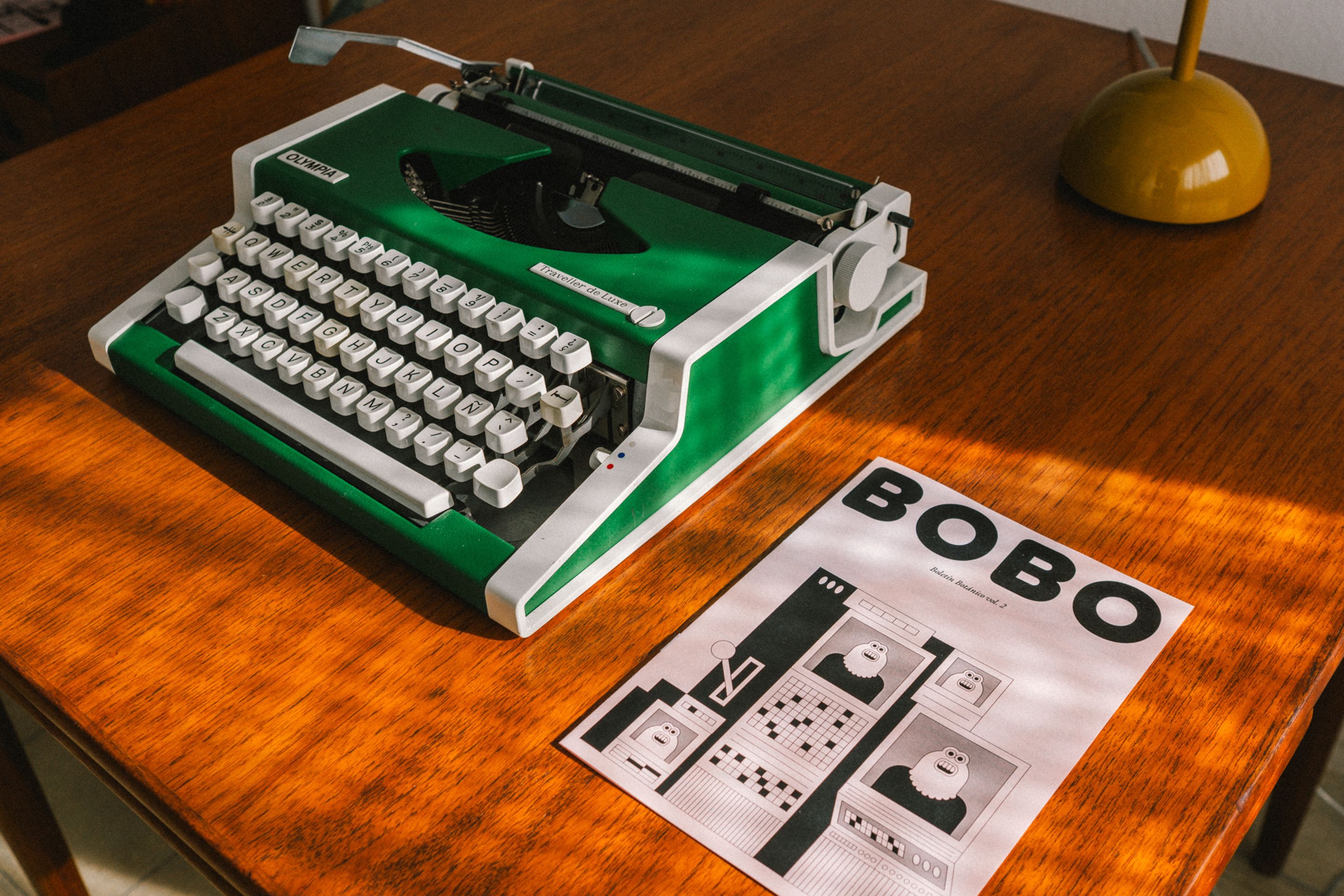 Máquina de escribir Olympia Traveller de Luxe de color verde vibrante sobre una mesa de madera, junto a un fanzine con el título «BOBO», disfrutando tumbado al sol.